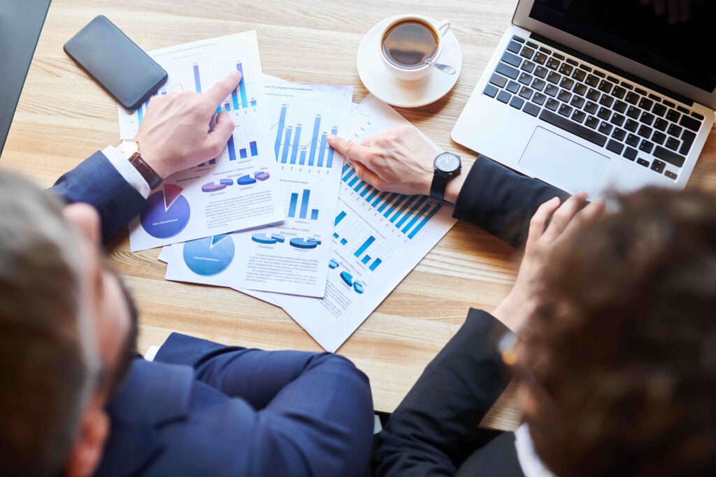 View of two young analysts sitting by table and pointing at financial charts while discussing statistics