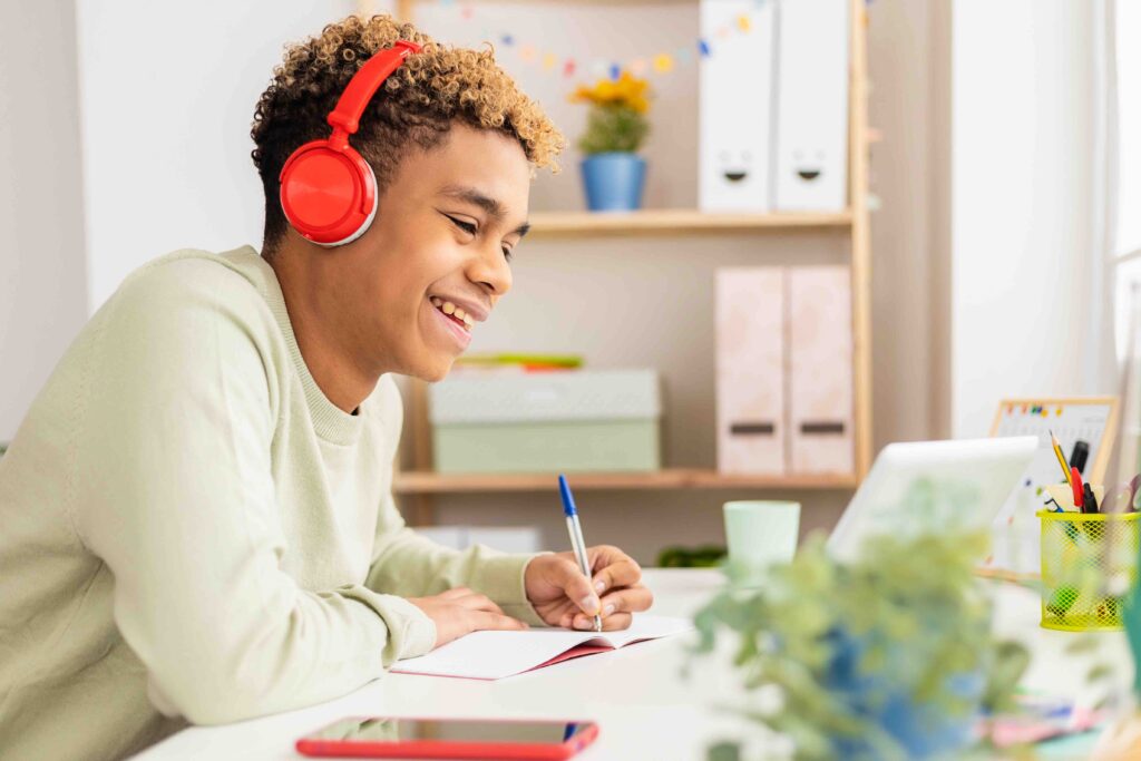 A young male student smiling while reviewing his school notes and listening to music. He is sitting down at his desk in an office setting with school supplies and notes around him.