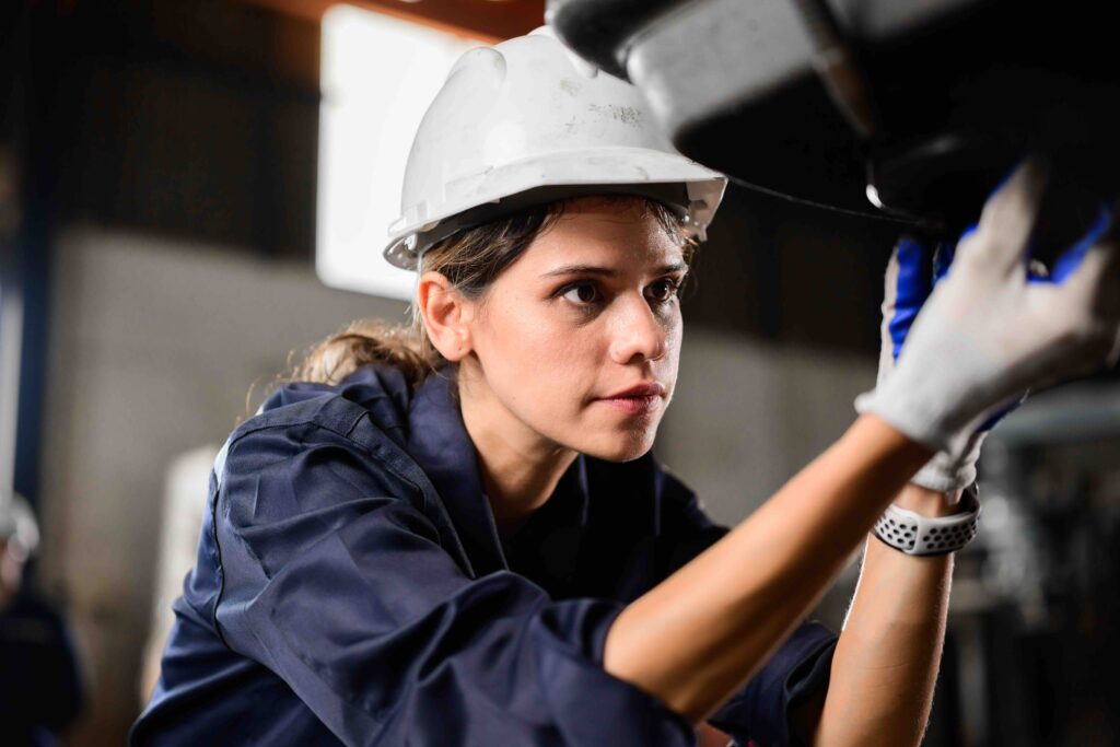 Young professional woman wearing a safety hat, focused on a machine, while working in a manufacturing plant