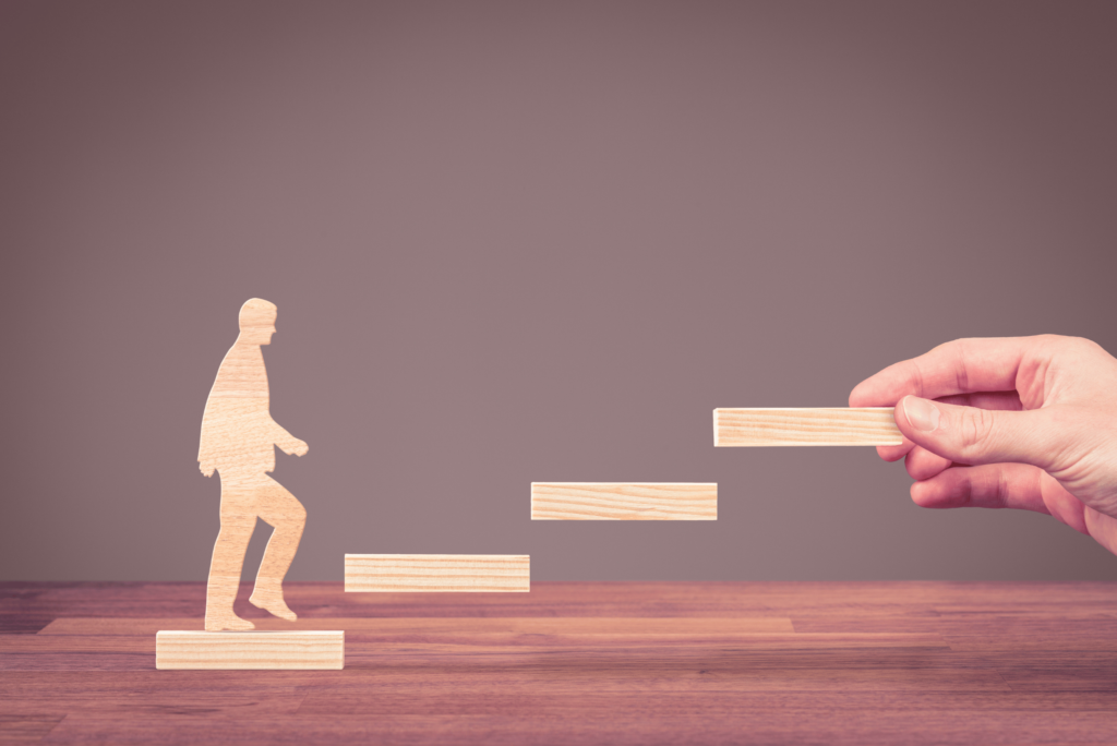 A man walking in an upward direction as a hand places wooden blocks for him to step on. This represents AJ Augur's consultation support in helping clients achieve career progression and development in plastics.